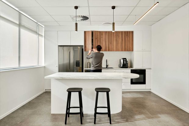 Bright contemporary office kitchenette with a large white island and two black bar stools in the foreground. The kitchen features white flat-panel cabinetry, a timber veneer overhead feature panel, integrated stainless-steel refrigerator, black oven, sink, coffee machine, and linear pendant lighting. Large windows provide abundant natural light, while the ceiling has exposed acoustic panels and services typical of a commercial workplace.