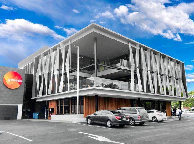 Exterior photograph of a contemporary multi-storey commercial building with a prominent glazed corner, white structural framing, vertical sculptural fins, timber-look cladding at ground level, and a projecting upper-floor canopy. Cars and a Genesis sign are visible in the forecourt under a bright blue sky.