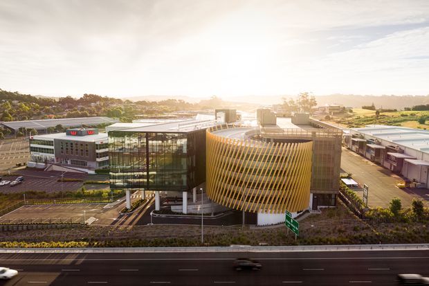 Aerial exterior view of a contemporary commercial complex beside a multilane highway. The main building features a glazed office or atrium volume paired with a prominent curved facade wrapped in closely spaced golden vertical fins. Additional low-rise commercial buildings, landscaped edges, parking areas, and surrounding suburban development are visible in the background.