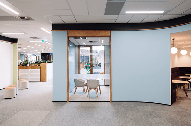 Contemporary commercial office interior with a light blue glazed meeting-room enclosure framed in warm timber, open-plan work areas, carpeted flooring, acoustic ceiling tiles, and a partially visible lounge or breakout area with pendant lighting. The image emphasizes the meeting room and surrounding office circulation space.