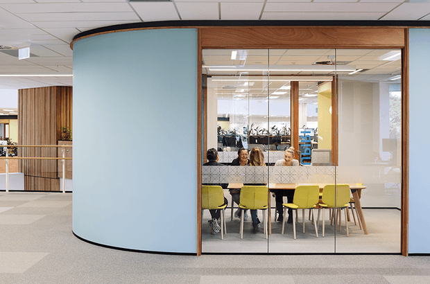 A contemporary commercial office interior with a glazed meeting room or collaboration room framed in warm timber. The room has a curved pale blue privacy wall, full-height glass partitions, a timber meeting table, and several bright yellow-green upholstered chairs. People are seated inside, with open-plan workspaces and timber feature screening visible beyond. The office has a suspended acoustic ceiling, carpeted flooring, and a bright, professional atmosphere.
