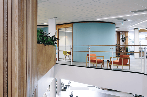 A contemporary commercial office interior viewed from an upper-level circulation area, overlooking a lounge and workspace. The scene features a curved pale blue partition, timber-clad walls and planter joinery, glass balustrades with warm timber handrails, exposed office glazing, orange lounge chairs, and a suspended grid ceiling with linear fluorescent lighting. The open void and layered floor levels create a bright, modern workplace atmosphere.
