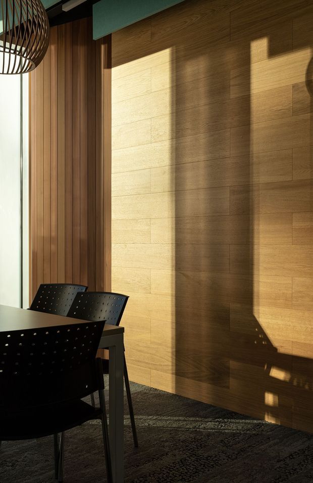 A warmly lit contemporary dining area with a dark dining table and perforated black chairs positioned beside full-height glazing. The room features a large fixed timber wall with horizontal panel joints, a vertical timber slat wall at the window, dark structural ceiling elements, and a sculptural open metal pendant light. Strong late-afternoon sunlight creates pronounced shadows across the timber surfaces and dark patterned floor rug.