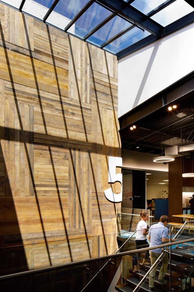 A multi-level commercial interior with a dramatic double-height atrium. A large wall is clad in reclaimed-looking timber boards arranged in varied horizontal and vertical sections, crossed by strong shadows from a glazed roof or skylight. Black steel beams, exposed services, glass balustrades, a stairway, contemporary pendant lighting, and work or dining areas create an industrial workplace or hospitality setting.
