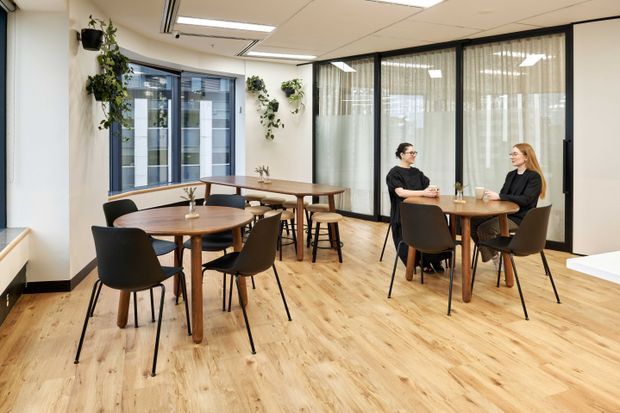 A bright contemporary office breakout and dining area with light timber-look flooring, several round and rectangular wooden tables, black chairs, pendant-free recessed ceiling lighting, and indoor climbing plants. Black-framed glazed meeting-room partitions with sheer curtains define the rear of the space, while large windows bring in daylight.