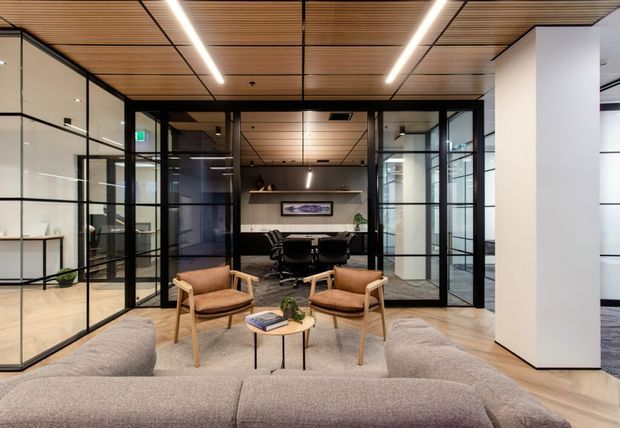 A refined contemporary office lounge and meeting area with a neutral upholstered sectional sofa, two timber-framed lounge chairs, and a small round coffee table in the foreground. Black-framed glass partitions enclose a meeting room with a conference table and chairs, while a timber slatted ceiling, linear recessed lighting, light timber flooring, and white walls create a warm, modern commercial interior.