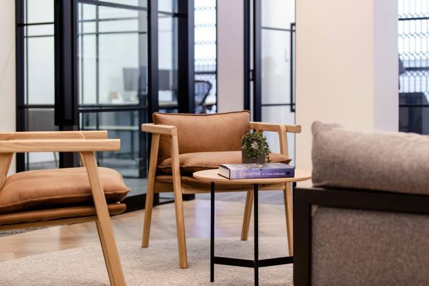 A refined contemporary living room with mid-century-inspired timber lounge chairs, tan leather upholstery, a small round black-framed side table, and a neutral upholstered sofa. Black-framed glazed partitions and a light timber floor create a bright, open interior with a calm, understated palette.