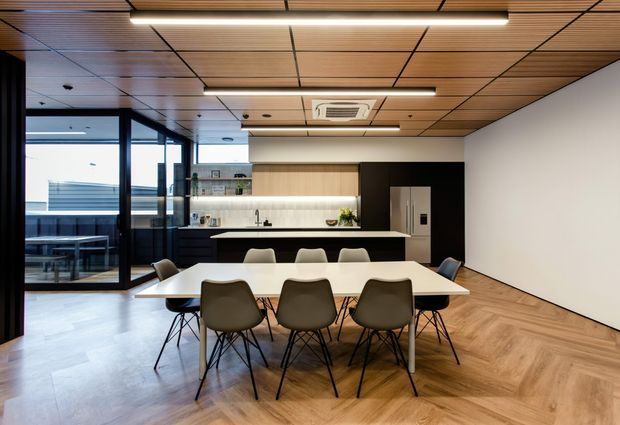 Contemporary open-plan kitchen and dining area with a long dining table, upholstered dining chairs, a linear kitchen island, integrated cabinetry, and a large refrigerator. The space features warm timber herringbone flooring, a timber slatted ceiling with recessed grid detailing, black-framed glazing, and a restrained black, white, and natural timber palette. Glazing opens toward an outdoor terrace or balcony.