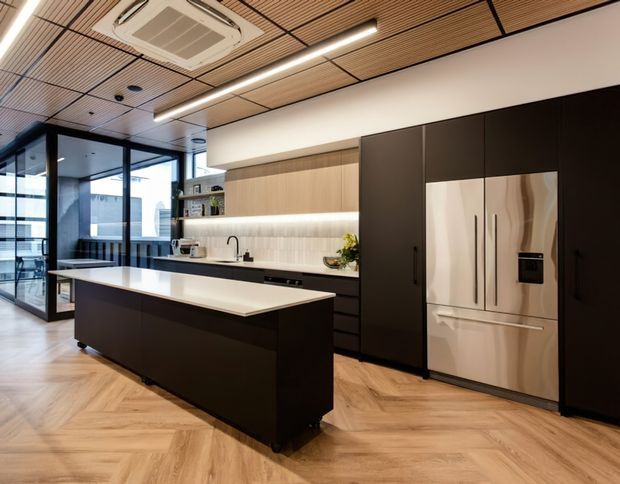 A contemporary kitchen with a large dark island, white waterfall benchtop, integrated appliances, extensive dark cabinetry, timber-toned upper cabinetry, and a tiled or slatted timber-look ceiling. Herringbone timber flooring, linear pendant lighting, and black-framed glazed partitions contribute to a refined modern office or residential kitchen setting.