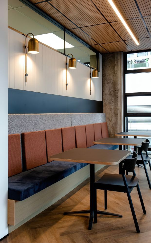 A contemporary commercial dining or hospitality interior with a built-in upholstered banquette, small dining tables, and black chairs. The space features warm timber flooring and a slatted timber ceiling, dark blue wall panelling, vertical light wall cladding, exposed concrete, black-framed glazing, and brass wall sconces.