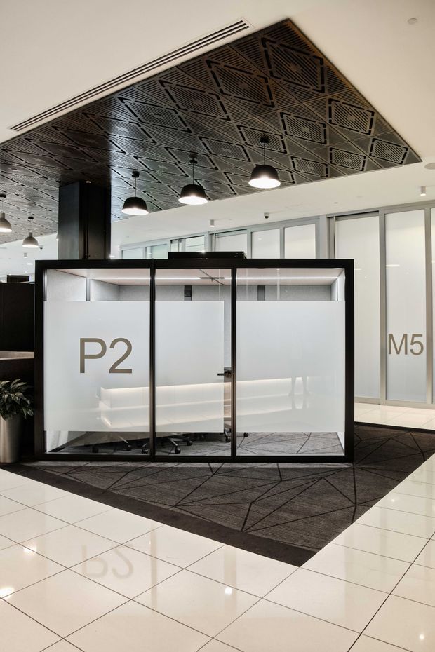 Modern commercial office interior showing a glazed meeting or work pod with frosted privacy panels labelled “P2” and “M5”. The space has a dramatic dark geometric perforated ceiling feature, suspended dome pendant lights, white tiled flooring, dark patterned carpet, black framing, and extensive frosted glass partitions.