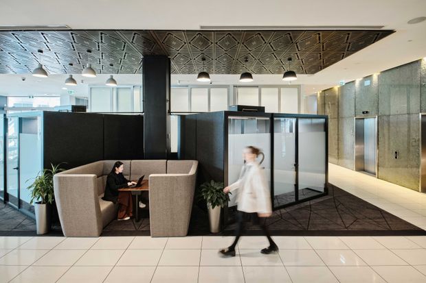 A contemporary commercial interior, likely a hotel or office lobby, with upholstered booth seating, black partition walls, glass-enclosed meeting spaces, a patterned metallic ceiling, pendant lighting, polished tiled flooring, and textured grey wall finishes. A blurred person walks through the foreground, while plants and soft furnishings add warmth to the otherwise refined, high-contrast space.