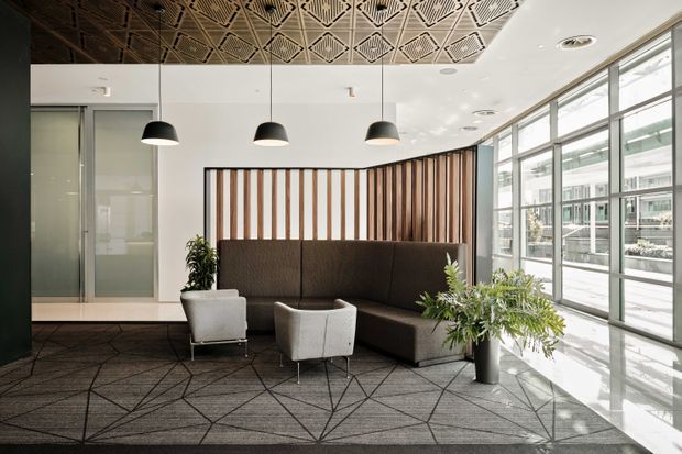 A bright contemporary commercial lounge or reception area with a dark sectional sofa, two white lounge chairs, pendant lights, indoor plants, floor-to-ceiling glazing, and a striking geometric patterned ceiling. A vertical timber slat feature wall frames the seating area, while patterned carpet defines the space.