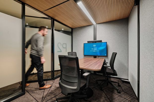 A compact contemporary meeting room or private office with a rectangular wood-finished conference table, several black ergonomic office chairs, a wall-mounted display, acoustic grey wall panels, a carpeted floor, black-framed glass partitions, and a slatted timber ceiling with linear lighting. A person is walking through the glazed entry area.