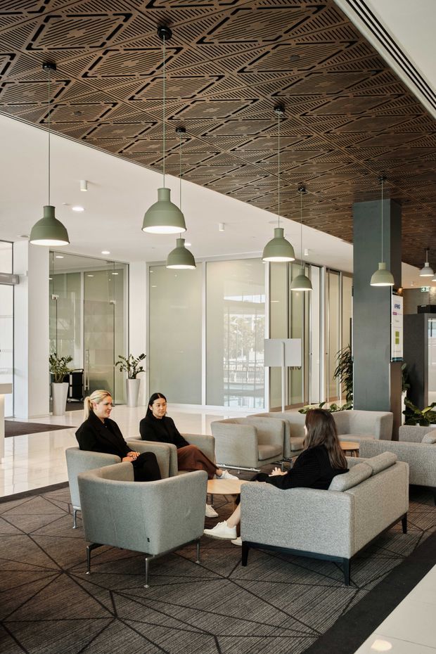 A contemporary commercial lobby or reception lounge with grouped upholstered armchairs and sofas around low coffee tables. The space features glazed partition walls, indoor plants, pendant lighting, polished light flooring, and a striking patterned timber acoustic ceiling.