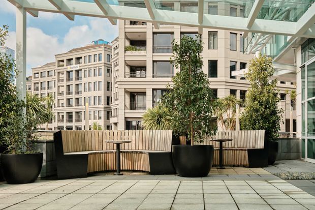 A landscaped urban rooftop terrace or elevated outdoor lounge with large square pavers, built-in timber bench seating, black pedestal tables, and oversized planters. A transparent glazed canopy and structural framing cover the terrace, while modern multi-storey apartment or commercial buildings surround the space.