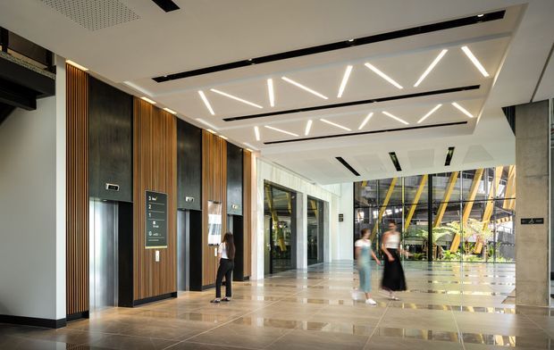 A spacious contemporary commercial lobby or building entry with polished stone flooring, elevator doors set into dark timber and black wall panels, and a high ceiling featuring linear recessed lighting. Full-height glazing opens the view toward a bright atrium with exposed structural elements. A few people are present in the background.