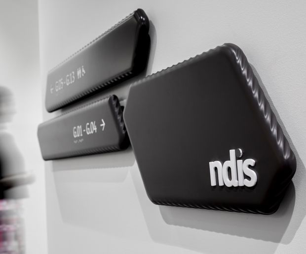 Close-up of three black, wall-mounted rectangular panels or display elements on a white wall. The foreground panel features raised white “ndis” lettering, while the two smaller panels behind it contain directional or technical text. The image has a monochromatic, graphic presentation with strong shadows and molded or padded-looking edges.