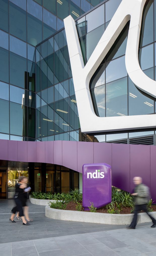 Street-level exterior view of a contemporary commercial building with a highly glazed curtain-wall facade, strong white sculptural framing around the windows, and a sweeping purple entrance canopy. A purple freestanding sign marks the NDIS building entrance, with landscaped raised planters and pedestrians in the foreground.
