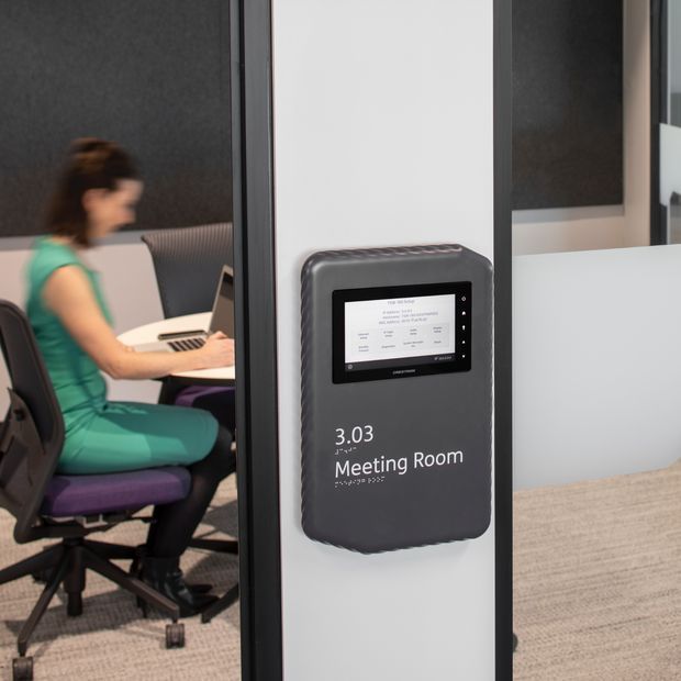 A modern office meeting room entrance with a wall-mounted digital room booking display labelled “3.03 Meeting Room.” Through the glass, a person works at a table in a furnished meeting room with office chairs, carpeting, and dark acoustic wall panels.