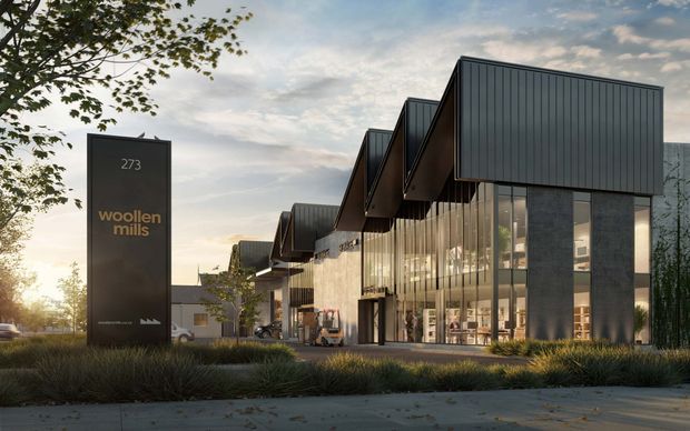 Exterior architectural view of a contemporary commercial development branded “woollen mills,” featuring a row of connected industrial-style retail or office buildings. The complex has dark metal-clad sawtooth roof forms, extensive floor-to-ceiling glazing, dark structural columns, landscaped planting, and a freestanding monument sign at the street edge, photographed in warm low-angle evening light.