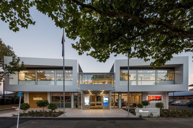 A contemporary two-storey commercial building viewed from the street, with a symmetrical white facade, large glazed office areas, a central recessed entrance, and a glazed connecting bridge at upper level. Signage and entry furnishings indicate a commercial office or business premises. Mature tree canopy and landscaped planting frame the frontage.