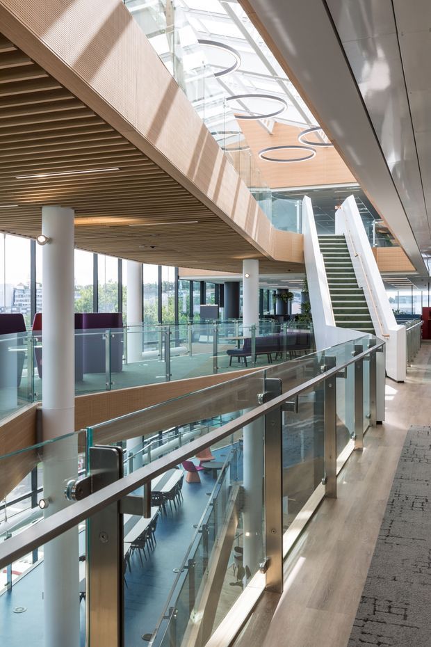 Bright multi-level contemporary commercial interior viewed from an upper-level circulation area, featuring a large glazed atrium, glass balustrades, a dramatic open stair, timber slatted ceilings, warm timber wall cladding, white structural columns, circular pendant lights, and extensive floor-to-ceiling glazing.