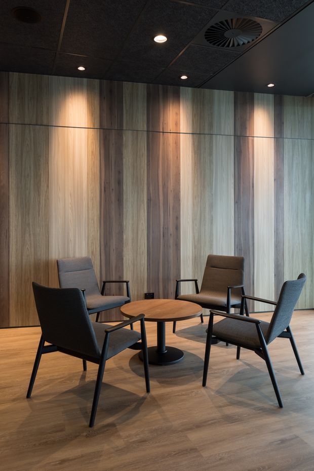 A dark, refined meeting or breakout area with four upholstered armchairs arranged around a round timber table. The room features full-height vertical timber wall panelling, matching timber-look flooring, a black acoustic tile ceiling with recessed downlights and exposed HVAC ventilation, creating a quiet contemporary commercial interior.