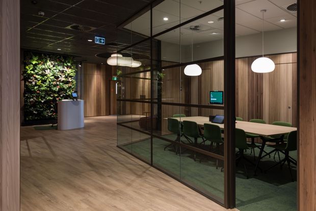 A contemporary commercial office interior featuring a glazed meeting room with a long light-coloured conference table, green upholstered chairs, and pendant lighting. The space has warm timber wall panelling, timber-look flooring, dark framed internal glazing, a reception desk, and a prominent living green wall.