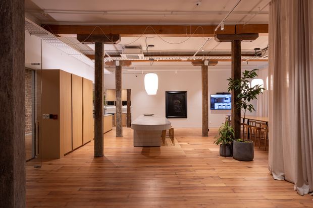 A warm, open-plan commercial interior with exposed timber posts and beams, polished timber flooring, concealed kitchen or service cabinetry along the left wall, and a central rounded island or reception counter. The space includes a dining or meeting area to the right, indoor plants, a framed wall display, track lighting, exposed ceiling services, and full-height curtains. The overall fit-out blends natural timber with a refined contemporary workspace or hospitality setting.