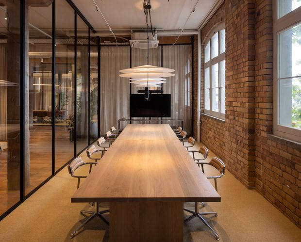 A warm, industrial-style meeting room with a long light-wood conference table surrounded by modern rolling office chairs. Exposed red-brown brick walls, black-framed glass partitions, high windows, a suspended projector, wall-mounted screen, exposed ceiling services, and layered pendant lights define the space.