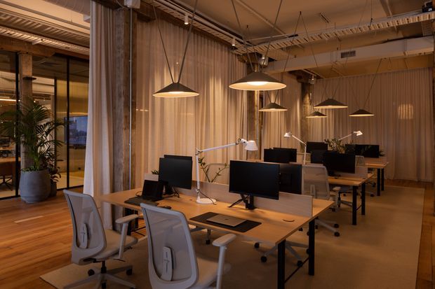 Warmly lit open-plan office with rows of timber workstations, black computer monitors, ergonomic mesh chairs, desk lamps, and suspended dome pendant lights. Exposed timber and metal ceiling services, vertical translucent curtains, glazed partitions, and indoor plants create a contemporary industrial workplace atmosphere.