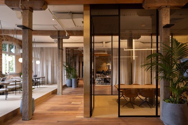 Open-plan commercial interior with exposed timber posts and beams, warm timber flooring, and a glazed meeting room enclosed by black-framed glass partitions. The space includes workstations and dining-style tables, abundant indoor planting, soft curtains, and contemporary linear pendant lighting. The design combines a warm, natural material palette with industrial structural character.