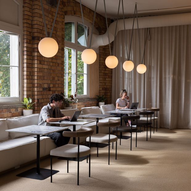 A warm, brick-lined coworking or café-style workspace with two people working at marble-topped tables. Tall industrial windows, exposed brick walls, suspended spherical pendant lights, built-in upholstered bench seating, freestanding chairs, and full-height curtains create a softly refined industrial interior. Large exposed ventilation ducts run near the ceiling.
