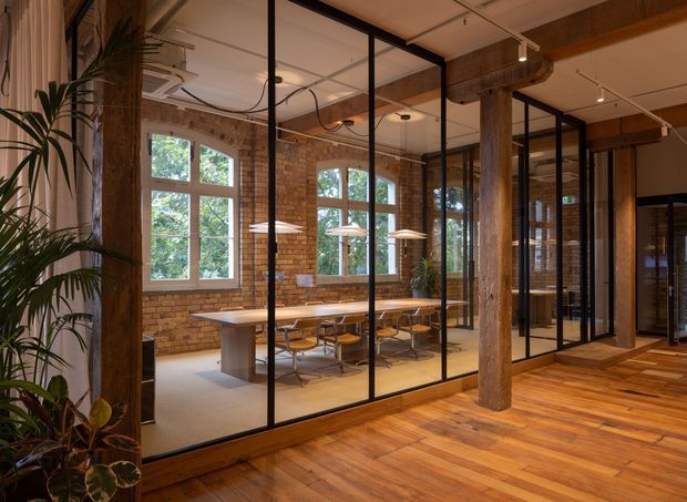 Interior view of a converted industrial-style workspace with exposed brick walls, substantial timber columns and beams, black-framed glass partitions, and a long communal meeting table. Warm timber flooring, pendant lights, abundant windows, and indoor plants create a refined yet organic office environment.