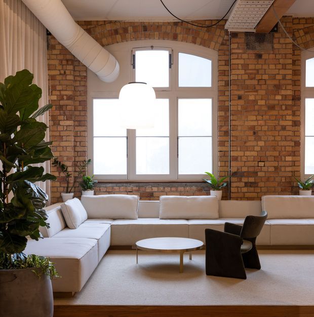 A bright loft-style living room with exposed warm brick walls, tall industrial windows, visible ductwork and a large white pendant light. A generous cream sectional sofa wraps around a low oval coffee table, with dark sculptural lounge chairs in front. Indoor plants soften the space, while the restrained palette and open, high-ceilinged composition create a refined industrial interior.