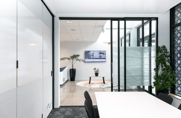 A bright contemporary office interior viewed from a meeting room. The foreground contains a large white meeting table with black chairs, opening through black-framed glass partitions to a reception or breakout area with a wall-mounted screen, indoor plants, polished flooring, and a patterned decorative screen along the right side. The space features clean white surfaces, dark framing, and abundant natural light.