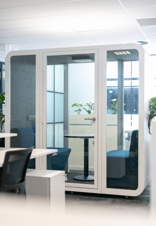 Bright contemporary office interior featuring a freestanding white-framed glass meeting booth or acoustic privacy pod with rounded corners. Blue upholstered seating and a small table are visible inside, with white desks and black office chairs in the foreground. The space has a white suspended grid ceiling, abundant glazing, and indoor plants.