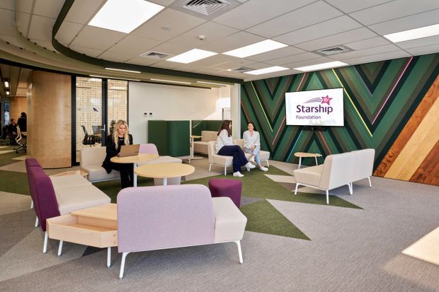 A bright contemporary office lounge and reception area with clustered pastel upholstered lounge chairs, small round timber tables, and a patterned carpet. The space features a dramatic geometric feature wall in green, black, pink, and timber tones, a suspended acoustic ceiling with recessed fluorescent lighting, and a Starship Foundation sign. Glass partitions and workspaces are visible in the background.