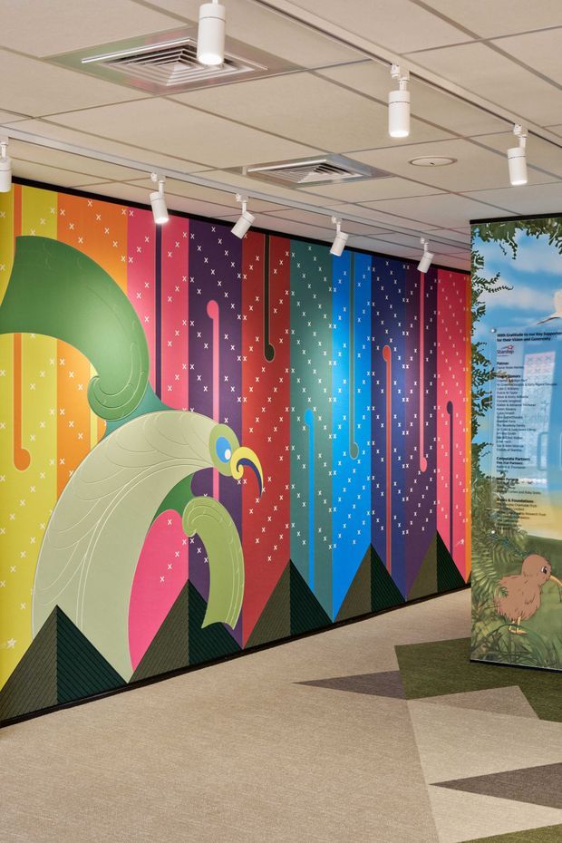 A brightly coloured commercial interior corridor or lobby featuring a large illustrated feature wall with stylised birds, multicoloured vertical panels, and geometric patterned carpet. Track spotlights and recessed air-conditioning are integrated into a suspended acoustic ceiling. A freestanding illustrated information panel is visible at the right.