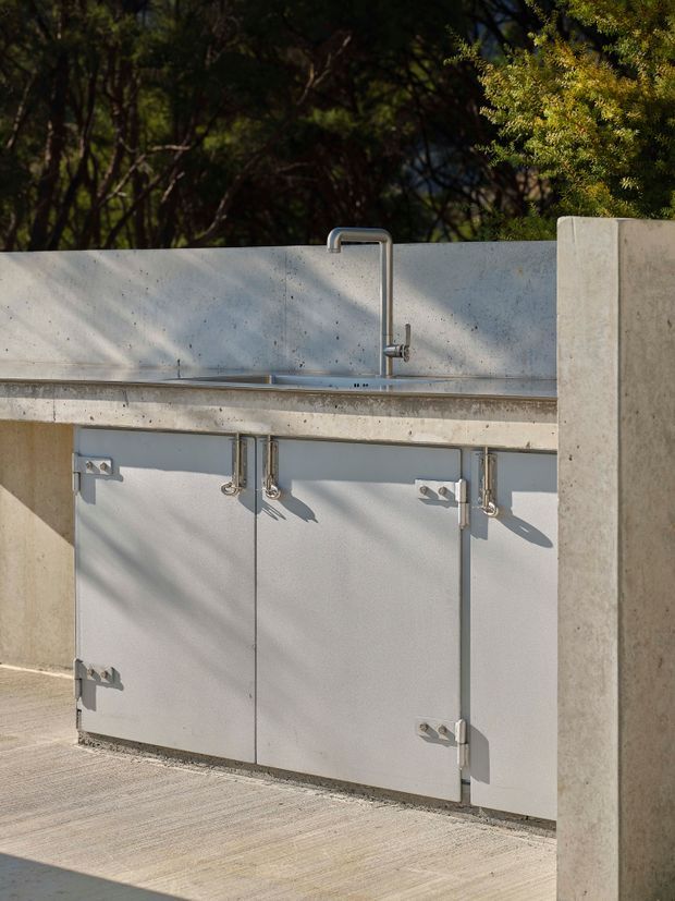 An outdoor concrete kitchen or utility station set against dense greenery. The installation features a pale grey concrete backsplash and countertop with an integrated stainless-steel sink and gooseneck tap, beneath which are simple light-grey cabinet doors with exposed metal hinges and hardware. A concrete side wall frames the right edge, and the surrounding paved surface and strong sunlight create a clean, utilitarian contemporary setting.