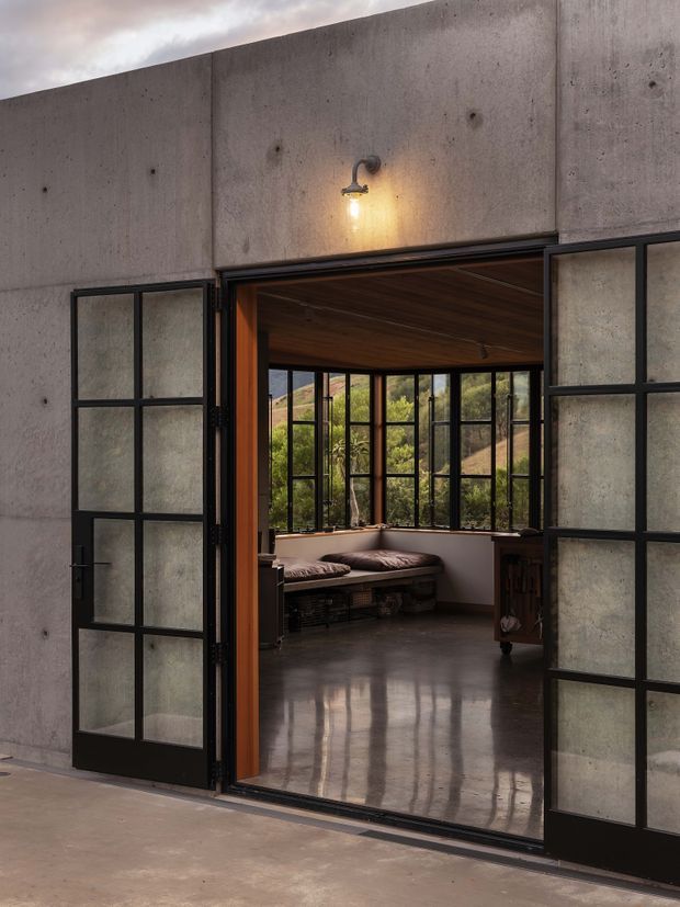 View through large black-framed glazed folding or pivot doors into a refined sunroom or lounge. The exterior facade is formed from exposed smooth concrete panels, with an exterior wall light above the opening. Inside, the room has a warm timber-lined ceiling, extensive black-framed glazing, a built-in cushioned bench, and a polished concrete floor. Vegetated hills are visible beyond the windows.