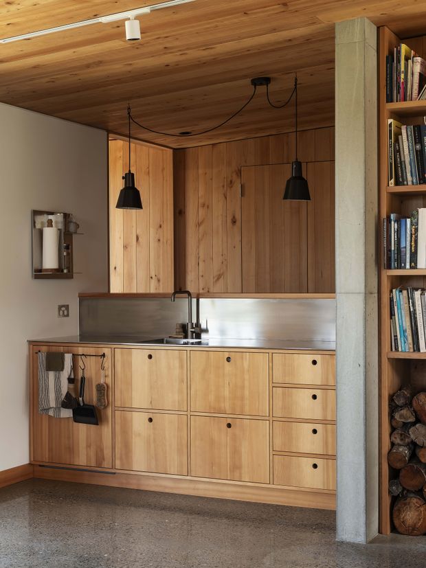 Compact, warm-toned kitchen with extensive light timber cabinetry and matching timber ceiling, a stainless-steel benchtop and splashback, and a small black sink and tap. Black pendant lights hang over the work area. A concrete structural column and open timber shelving with books and stacked firewood frame the kitchen, while the floor appears to be polished concrete or terrazzo.