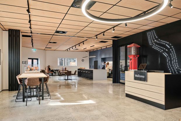 A contemporary commercial reception and dining area with a polished concrete floor, light timber slatted ceiling, black feature walls, track lighting, and a light timber reception desk. The space includes communal dining tables and chairs, a service counter or bar, and a distinctive red vintage-style telephone booth display beside the reception area. A black wall features a curved dotted light installation and Waitomo signage.