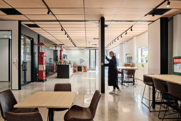 A bright contemporary commercial interior with an open-plan café or hospitality-style breakout area. The space features polished concrete or terrazzo flooring, timber-look acoustic ceiling panels with exposed grid lines, black structural columns and track lighting, extensive internal glazing, dining tables, stools and upholstered chairs. Red vintage-style display elements and a service counter add visual accents, while a person is shown moving a black-framed sliding or pivoting partition.
