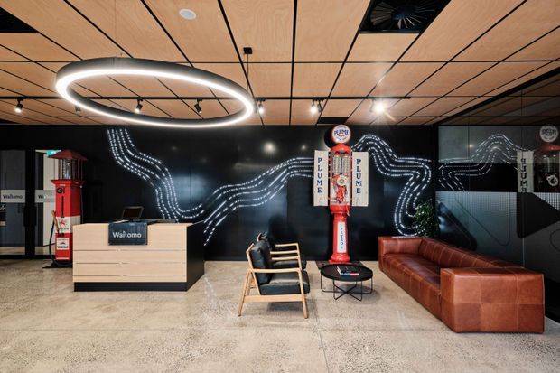 Contemporary commercial reception and lounge area with a timber-panelled ceiling, polished concrete floor, black feature wall with illuminated wave-like graphics, circular pendant lighting, a pale timber reception desk, vintage petrol pumps, a leather sofa, and timber-framed lounge chairs.