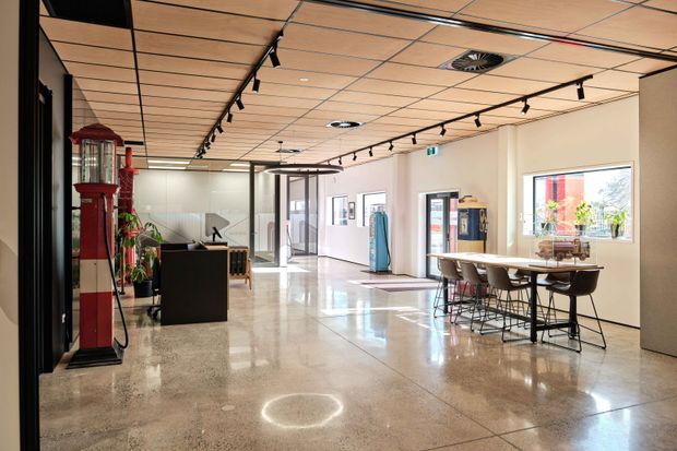 Bright contemporary commercial interior with an open reception and shared breakout area. The space features polished concrete flooring, a timber-panelled suspended ceiling, white walls, black track lighting, glass partitions and doors, a reception counter, and a long communal table with bar stools. A red vintage-style telephone booth or display structure provides a strong accent along the left wall.
