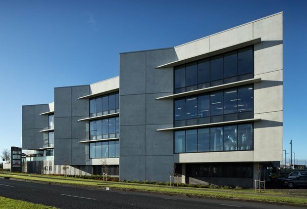 A contemporary multi-storey commercial building with a geometric facade composed of large grey panels, broad dark-framed glazed windows, and projecting horizontal sunshade ledges. The building fronts a paved road with minimal landscaping and parked vehicles, under a clear blue sky.
