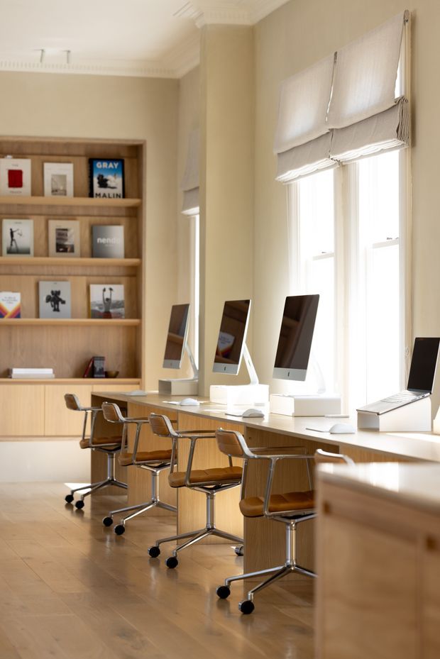 A warm, light-filled workspace or studio with a long timber workbench running along tall windows, several desktop computers, and brown leather swivel chairs. Built-in timber shelving displays books and framed prints, while cream Roman blinds and decorative ceiling cornices add a refined traditional touch. The room has pale timber flooring and a calm, curated atmosphere.