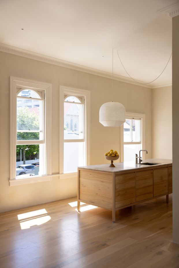 A bright, refined kitchen in a period-style room, featuring a freestanding timber island with integrated drawers, a pale stone benchtop, undermount sink and dark tapware. Tall arched-top windows with decorative mouldings, ornate ceiling cornices and timber flooring give the space a heritage character, while the simple white pendant and pared-back cabinetry add contemporary contrast.